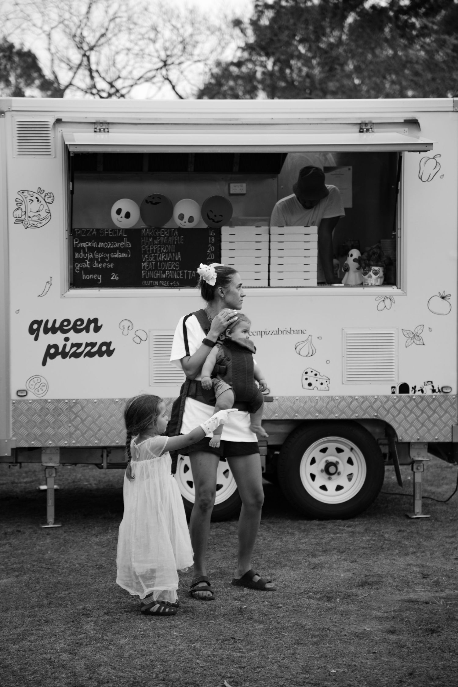 Black and white photo of mother with baby and young girl at Queen Pizza food truck at outdoor event.