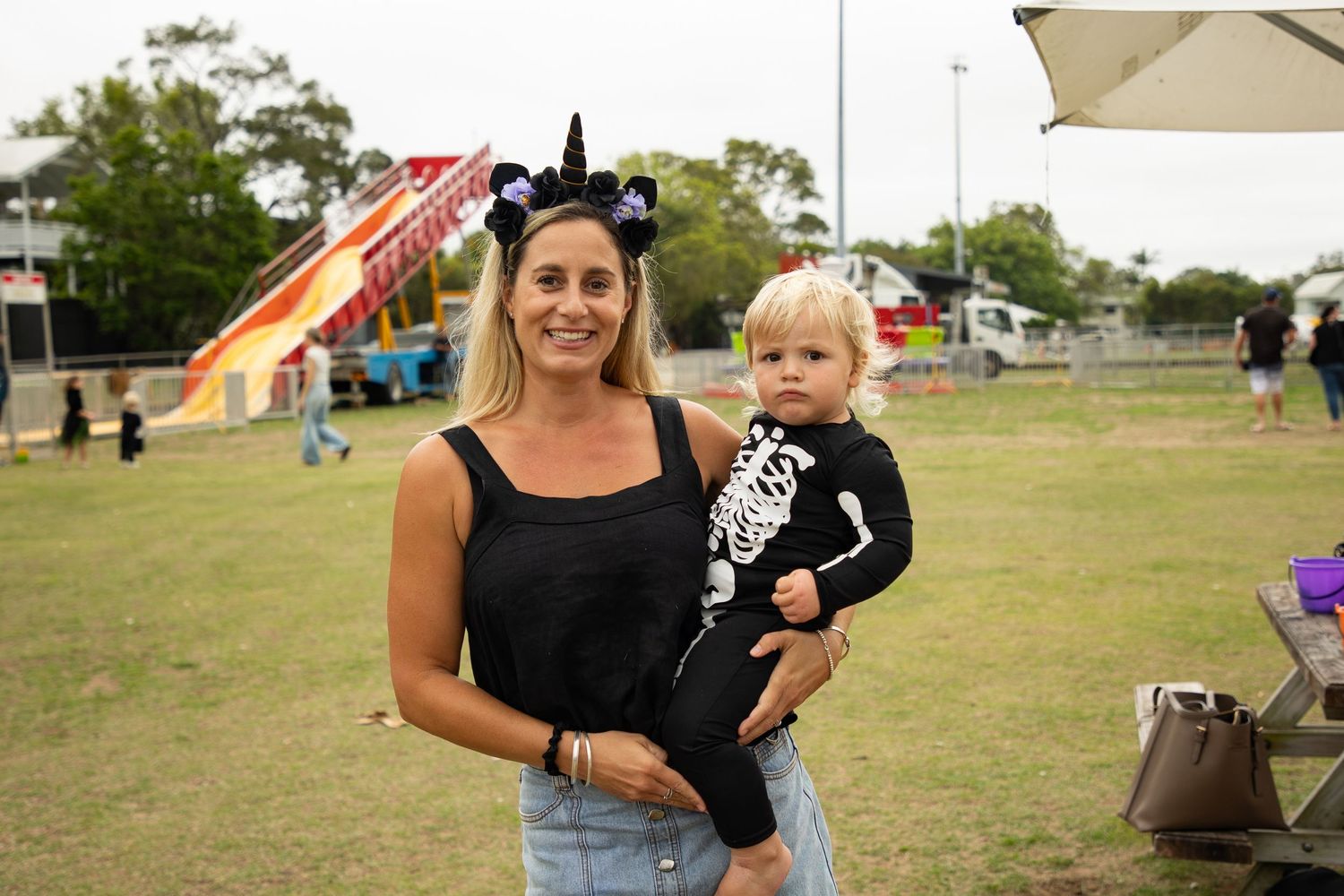 Woman holding toddler in skeleton Halloween costume at outdoor festival with carnival rides in background.