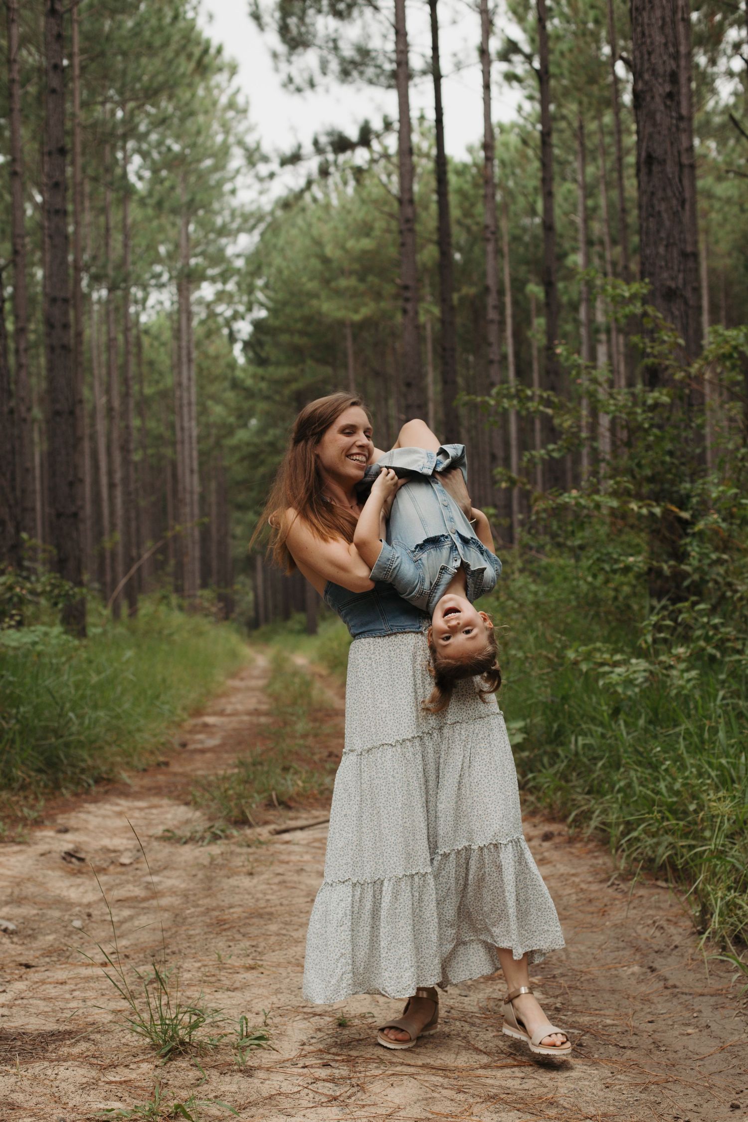 Mother playfully holding child upside down while laughing on a pine forest trail.