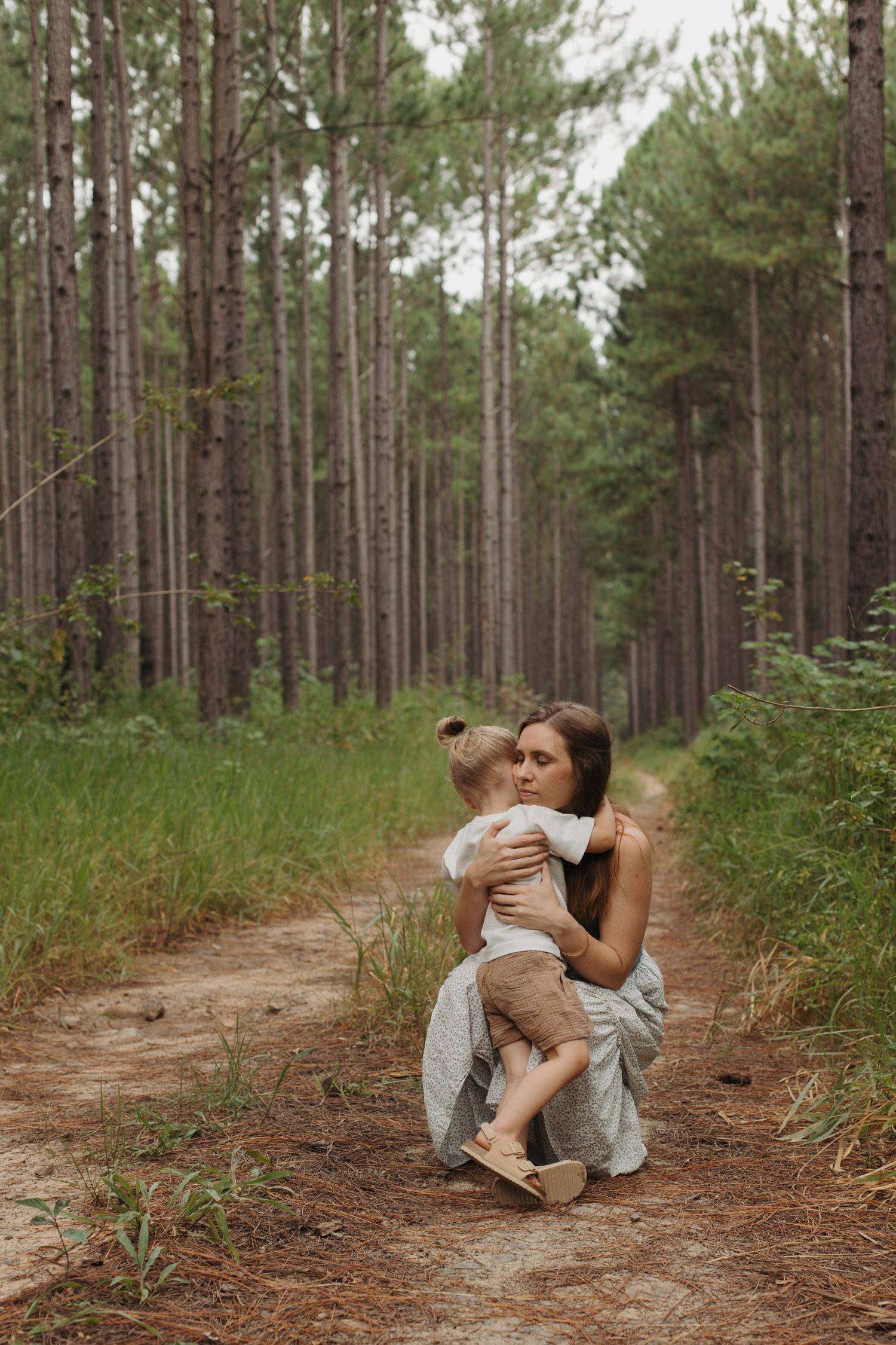 Mother embracing toddler on a forest trail surrounded by pine trees.