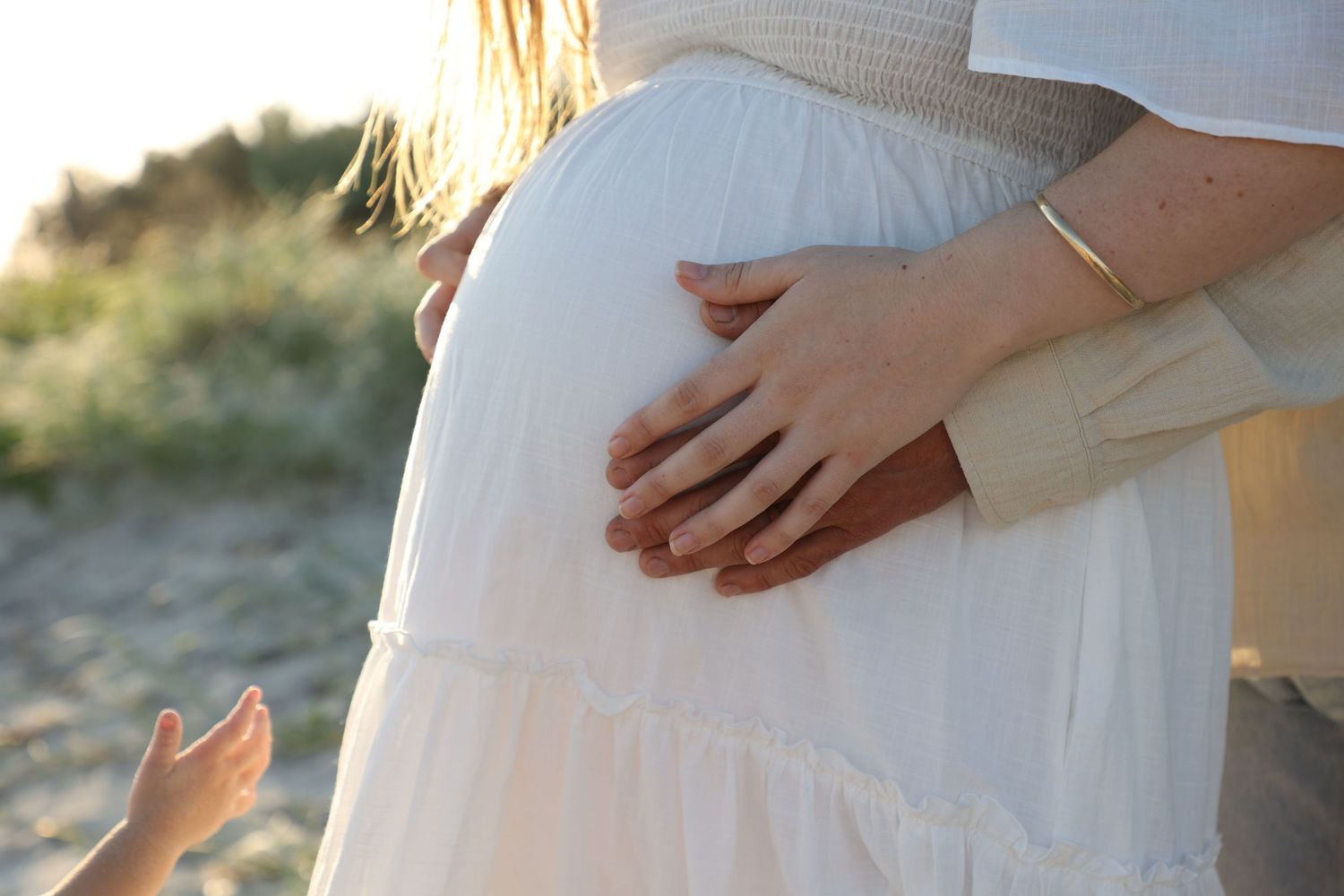 Close-up maternity photo on the beach with mother holding baby bump beside partner.