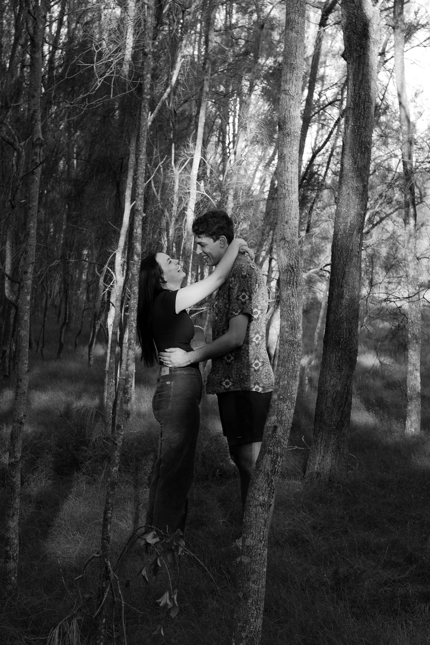 Black and white photo of romantic couple embracing between trees in a forest setting.