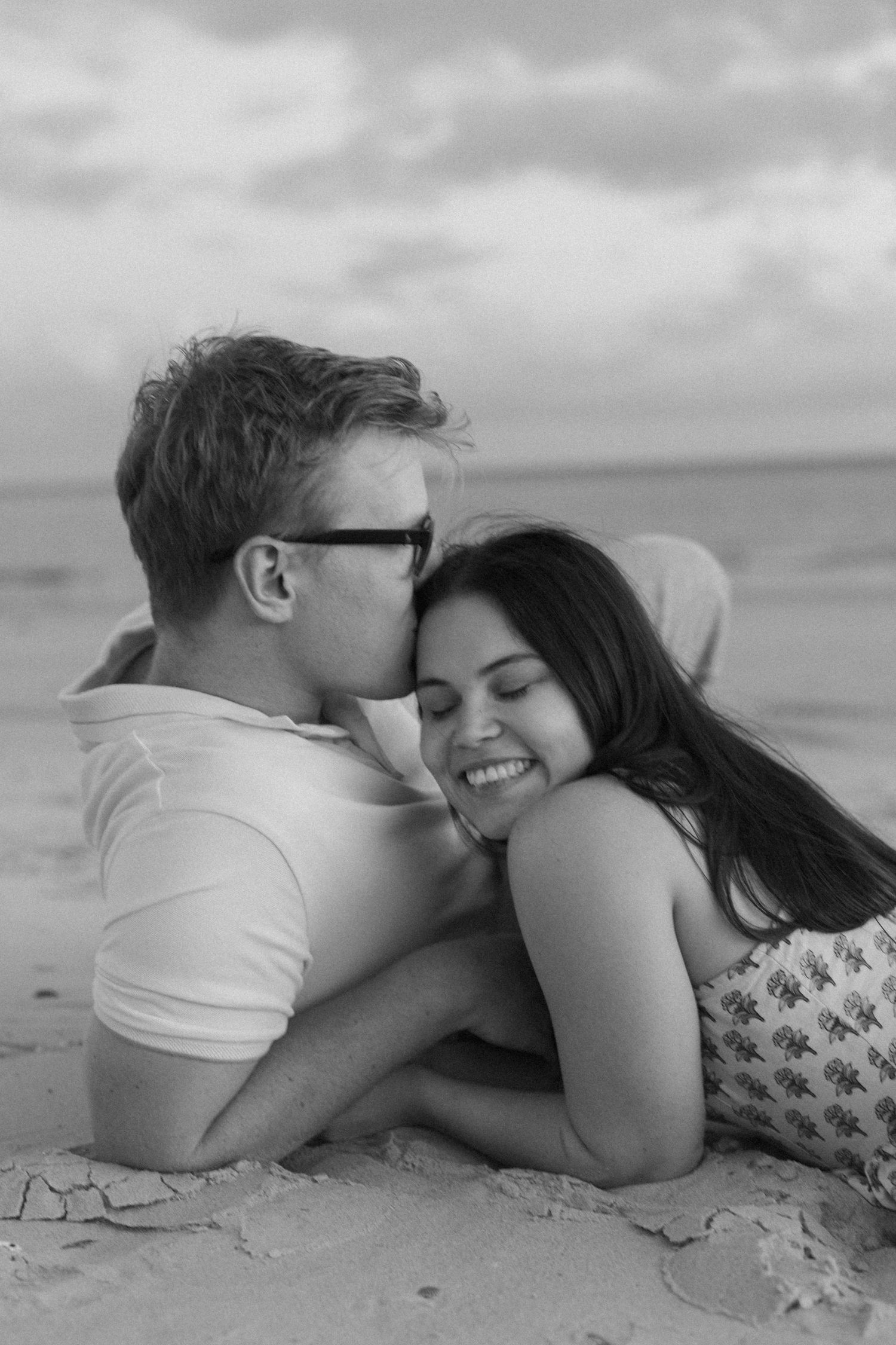Black and white photo of a couple lying on beach sand, man kissing womans cheek as she smiles joyfully.