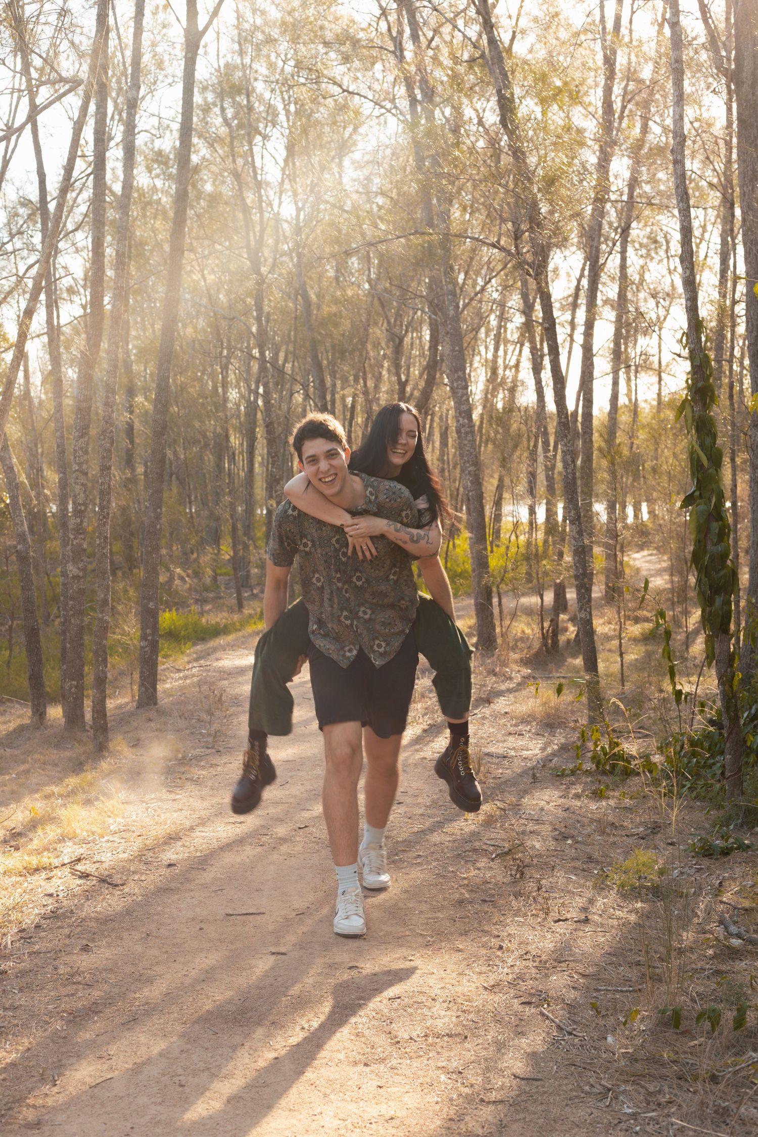 Man giving woman a piggyback ride down a sunny forest trail bathed in warm golden light.