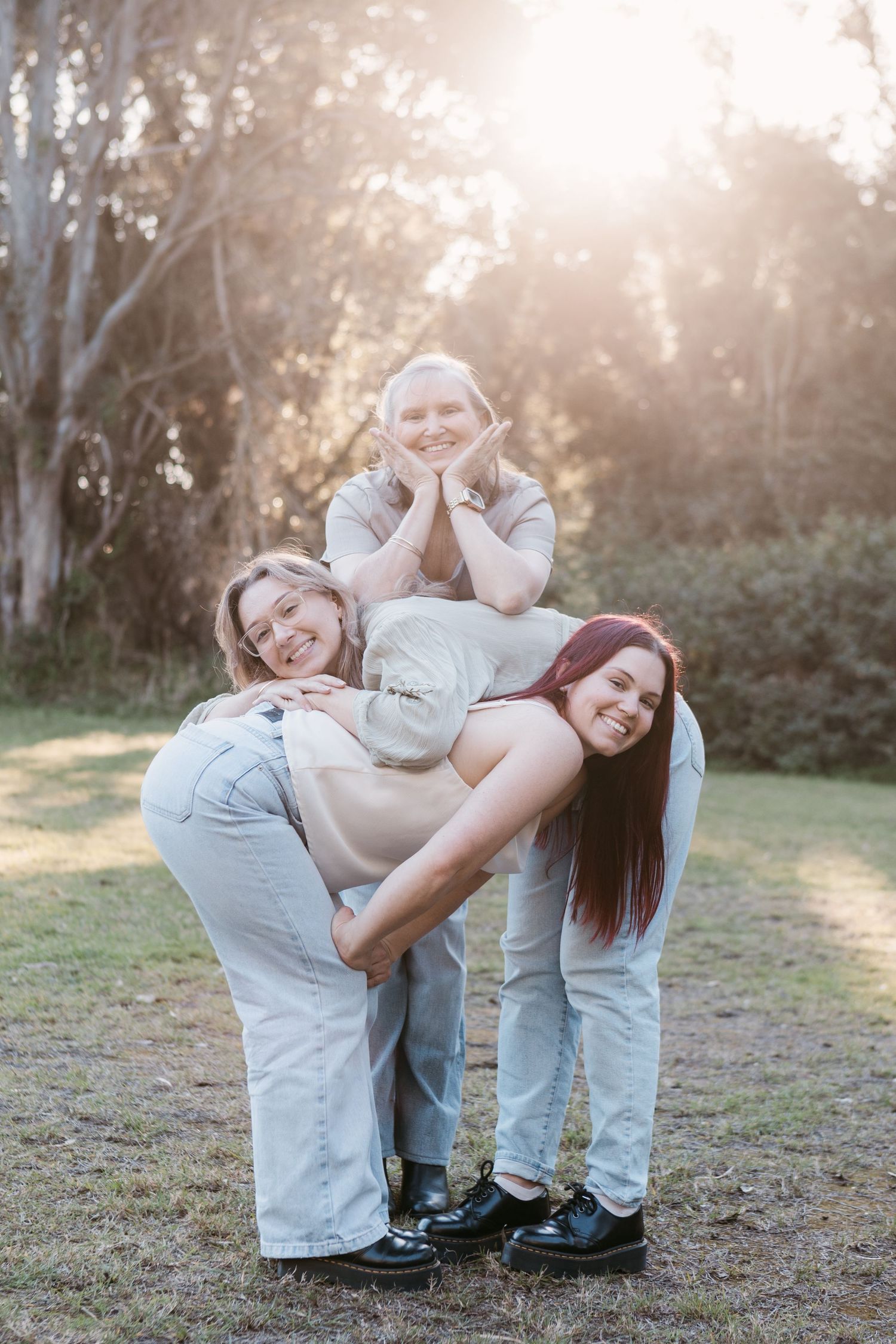 Three women posing playfully outdoors in golden sunlight, smiling and stacking in a fun pyramid formation.