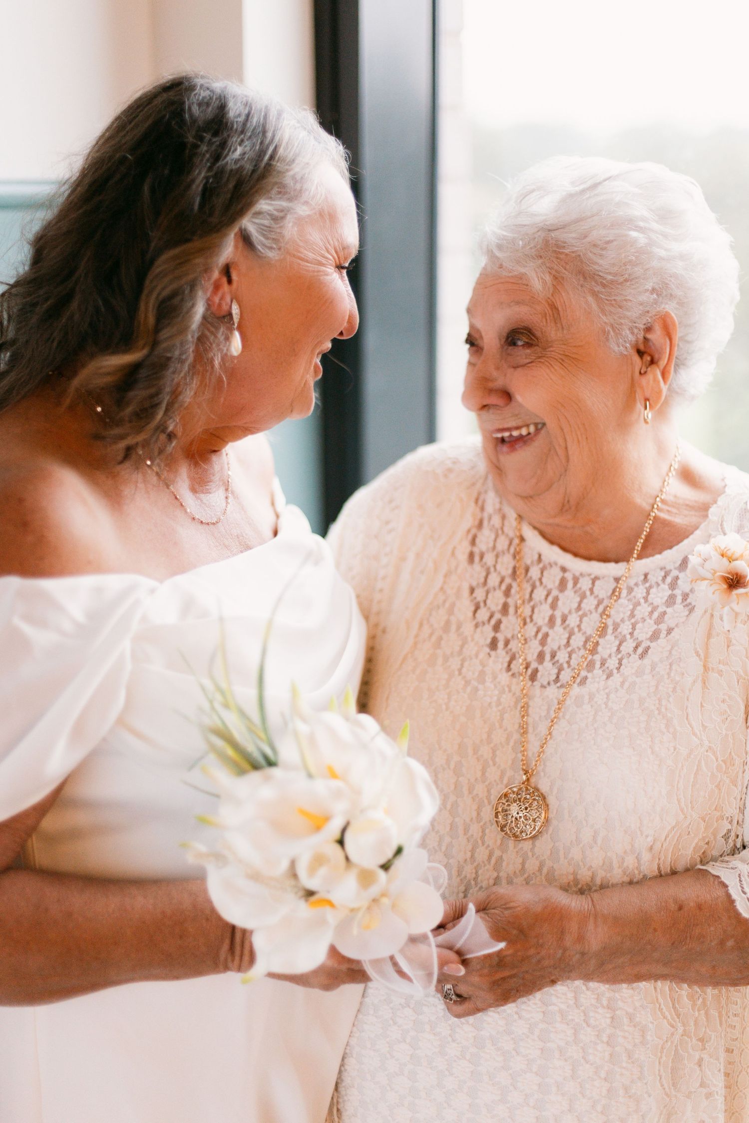 Mature bride in white gown holding flowers, sharing a joyful moment with elderly woman.