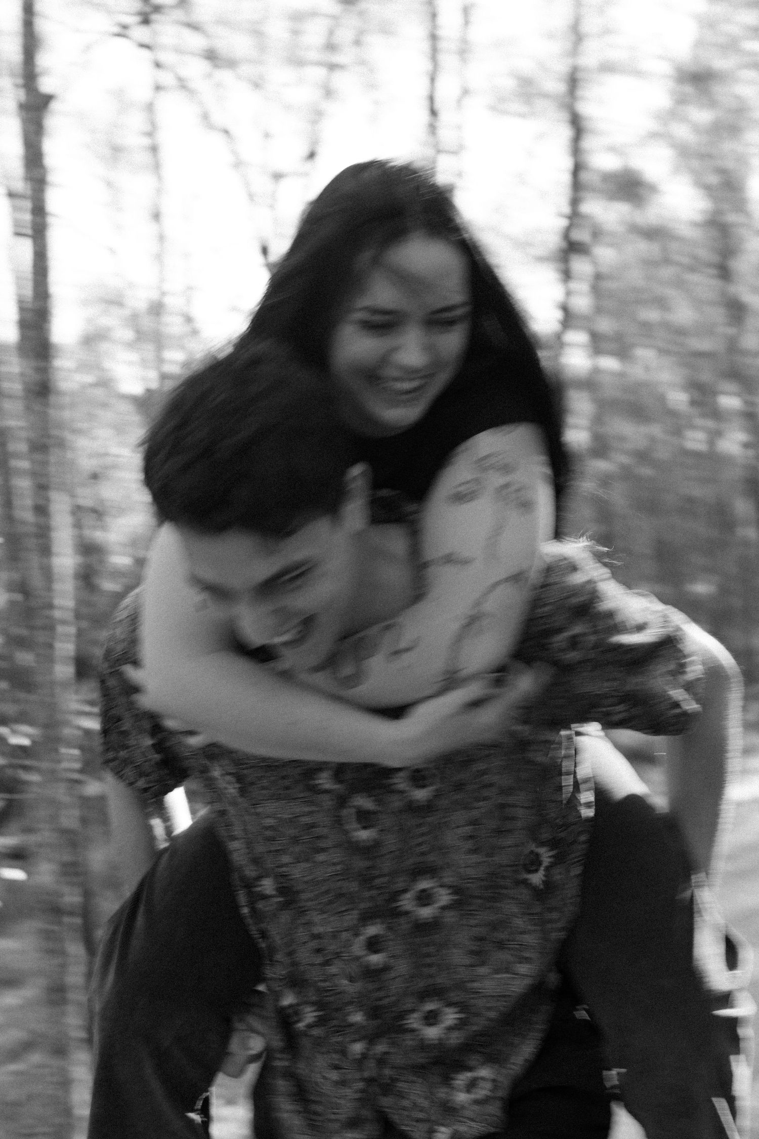 Black and white photo of laughing couple doing a piggyback ride in a forest setting.