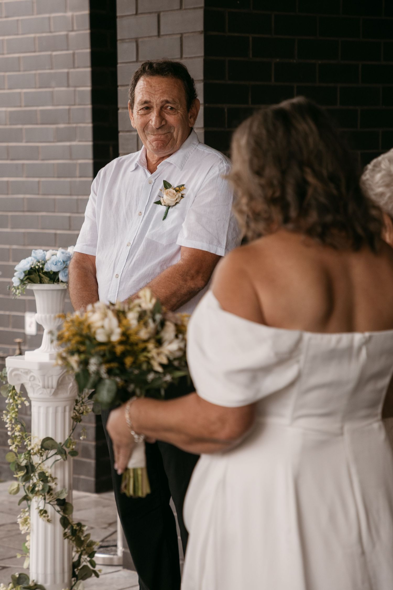 Groom with boutonniere smiling at bride in white off-shoulder dress holding floral bouquet at wedding ceremony.