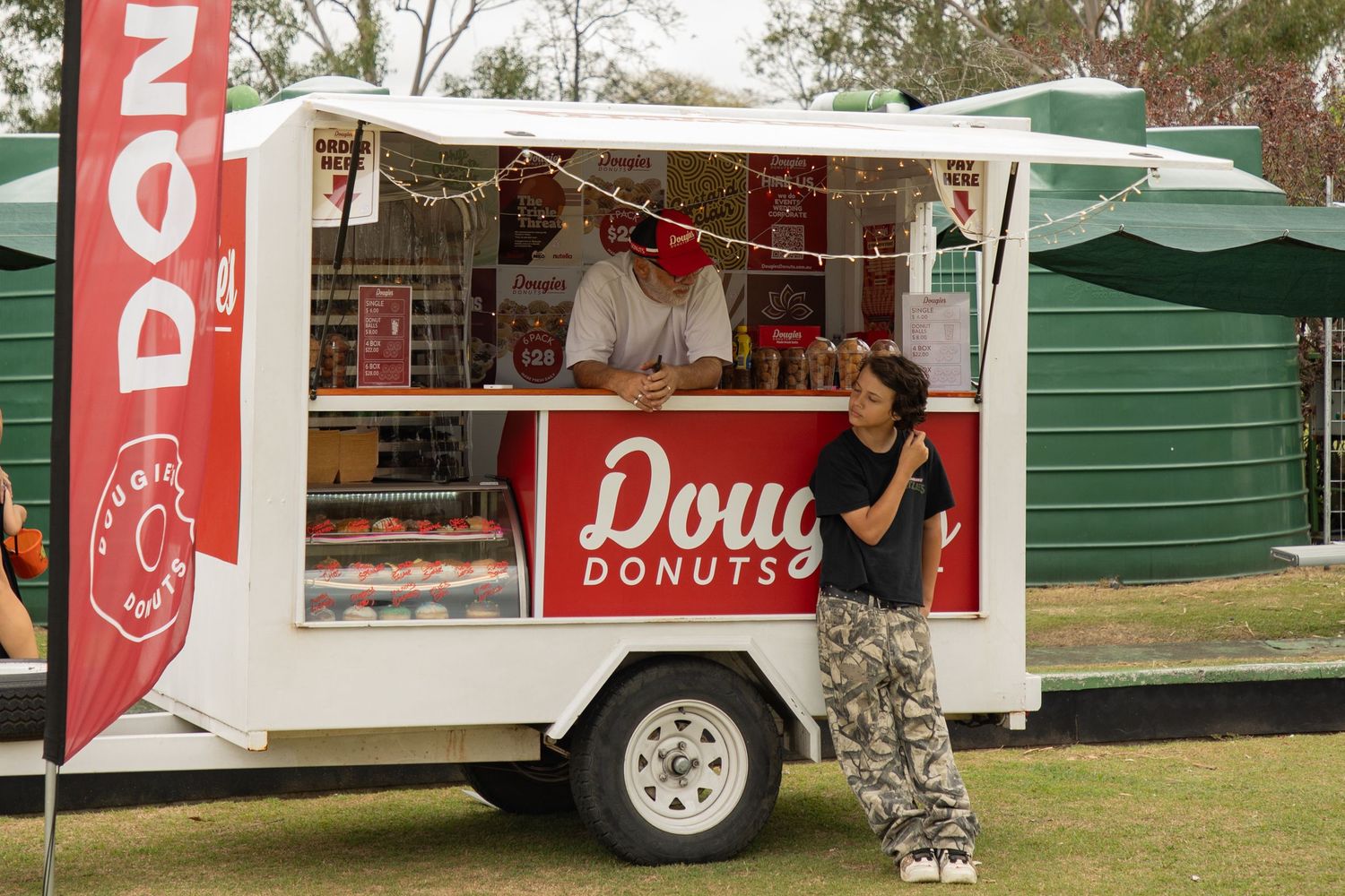 A man stands beside a Doughies Donuts food trailer at an outdoor event on a sunny day.