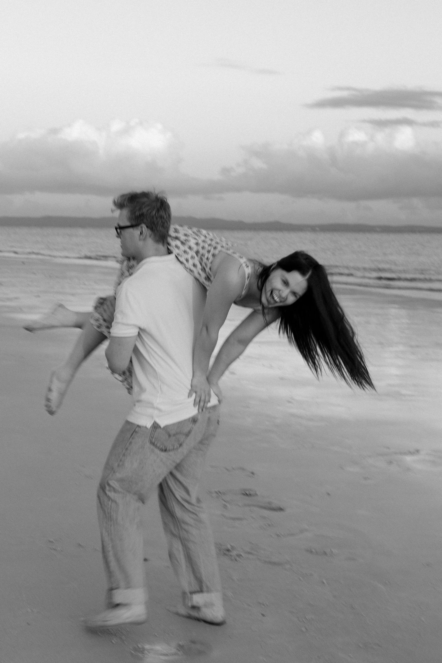 Black and white photo of a man playfully carrying a laughing woman on his back along a sandy beach.