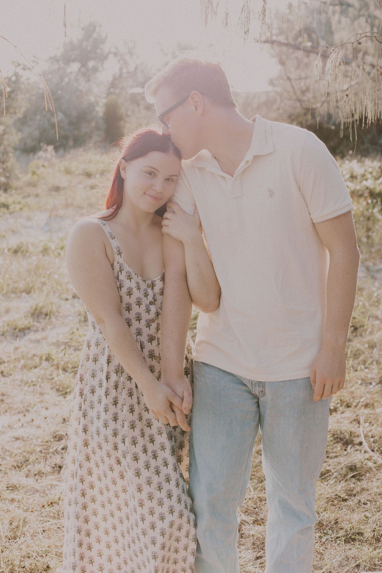 Couple holding hands in a sunlit field, man whispering to woman in floral dress.