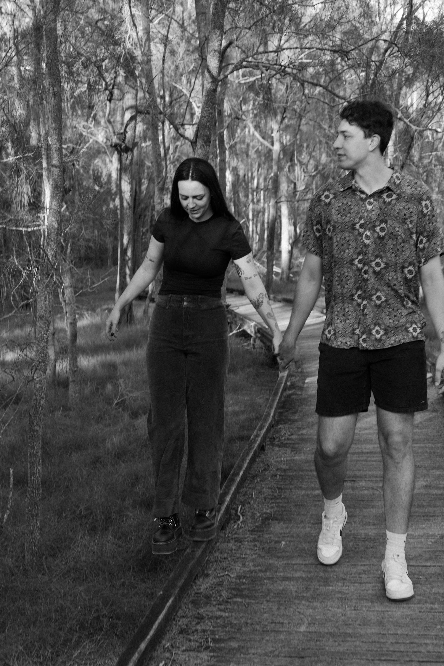 Black and white photo of couple holding hands while woman balances on a wooden boardwalk rail in forest.