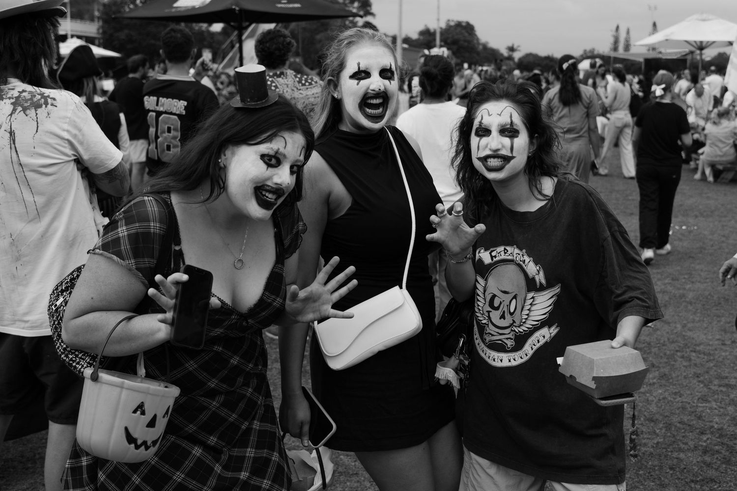 Three women in Halloween clown face paint pose together at an outdoor festival in black and white.