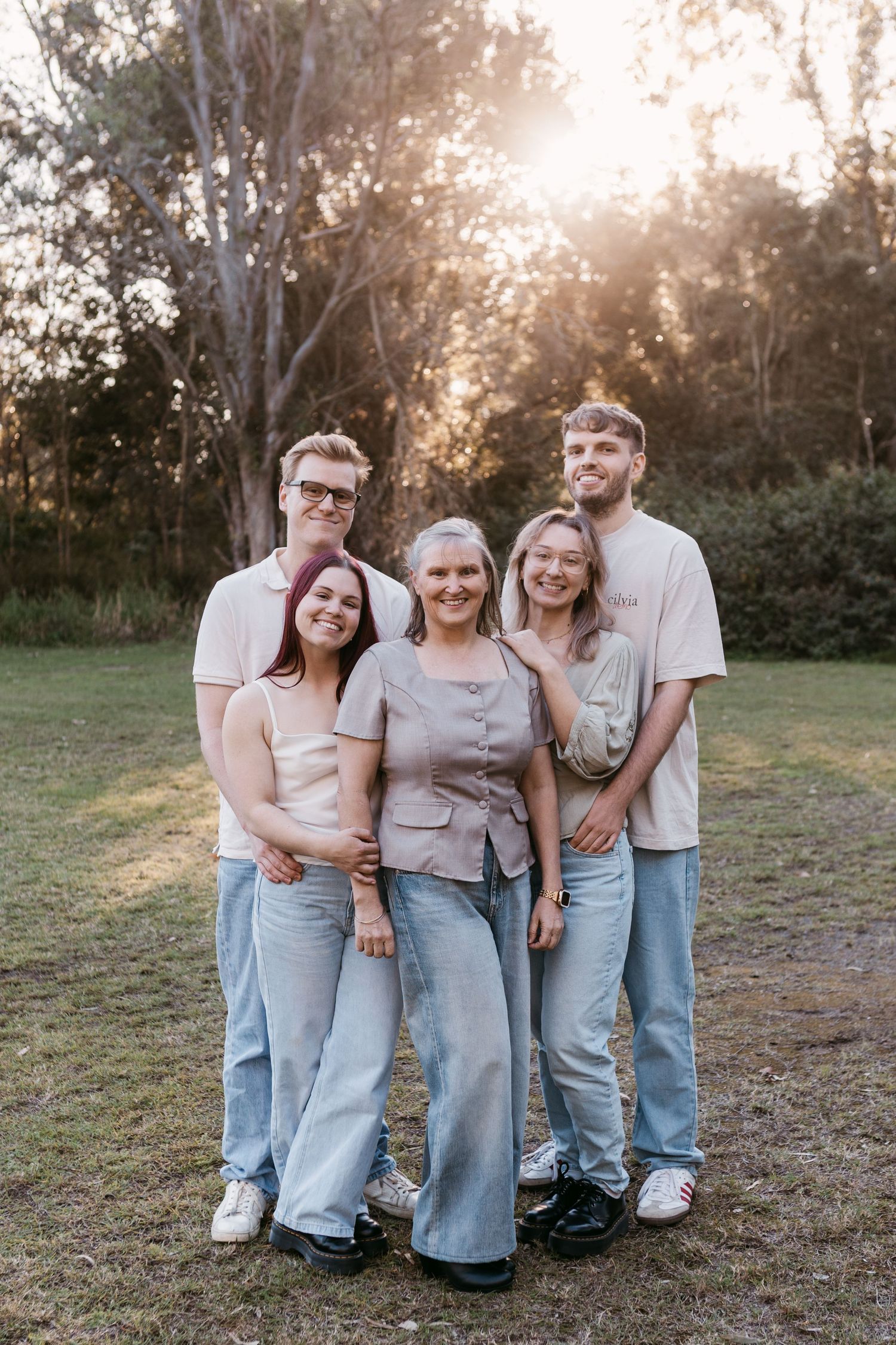 Family of five smiling together outdoors at golden hour, wearing casual jeans and neutral tones in a park setting.
