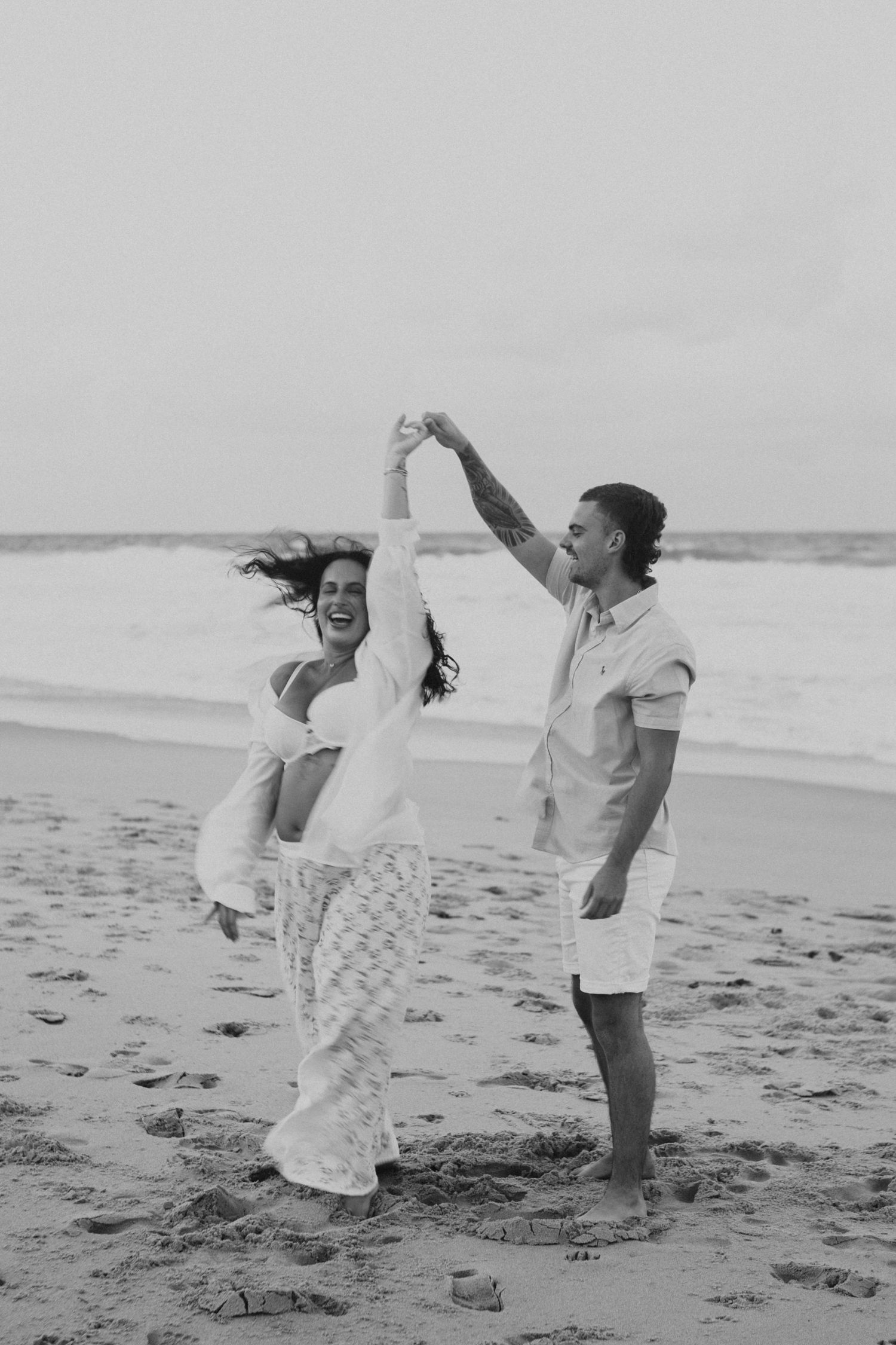 Black and white photo of a joyful couple dancing on a beach, woman laughing as man spins her near ocean waves.