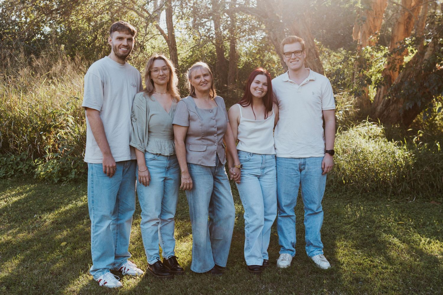 Family of five posing outdoors in matching jeans and neutral tops, smiling in a sunlit green park setting.