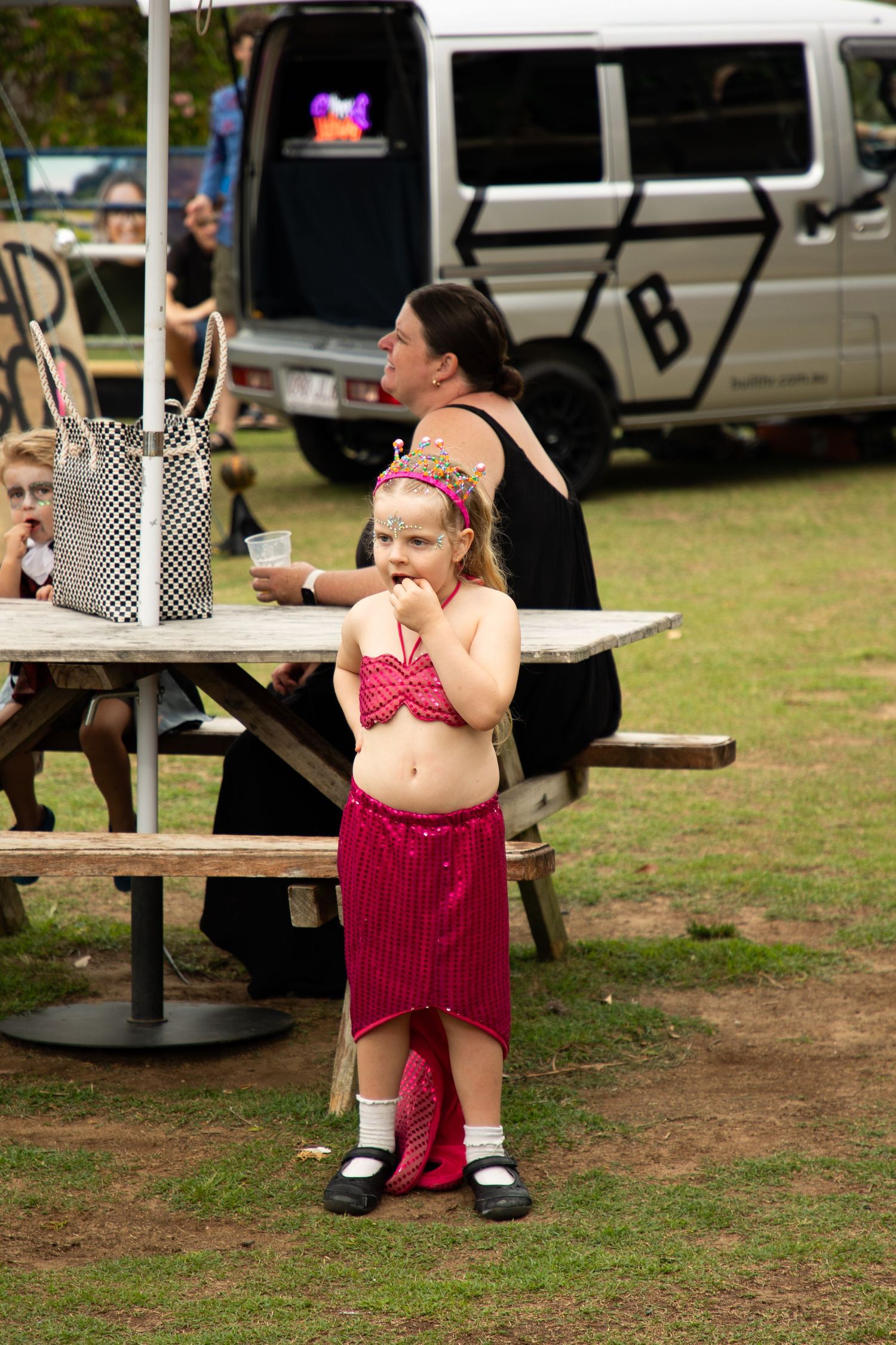 Young girl in pink mermaid Halloween costume stands at outdoor festival picnic area.