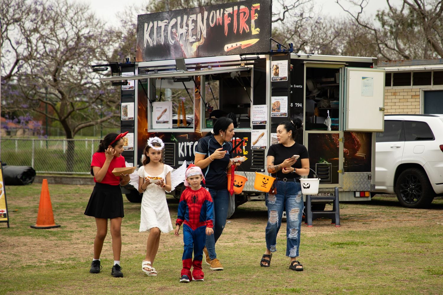 Family group in Halloween costumes pose together near Kitchen on Fire food truck at outdoor event.