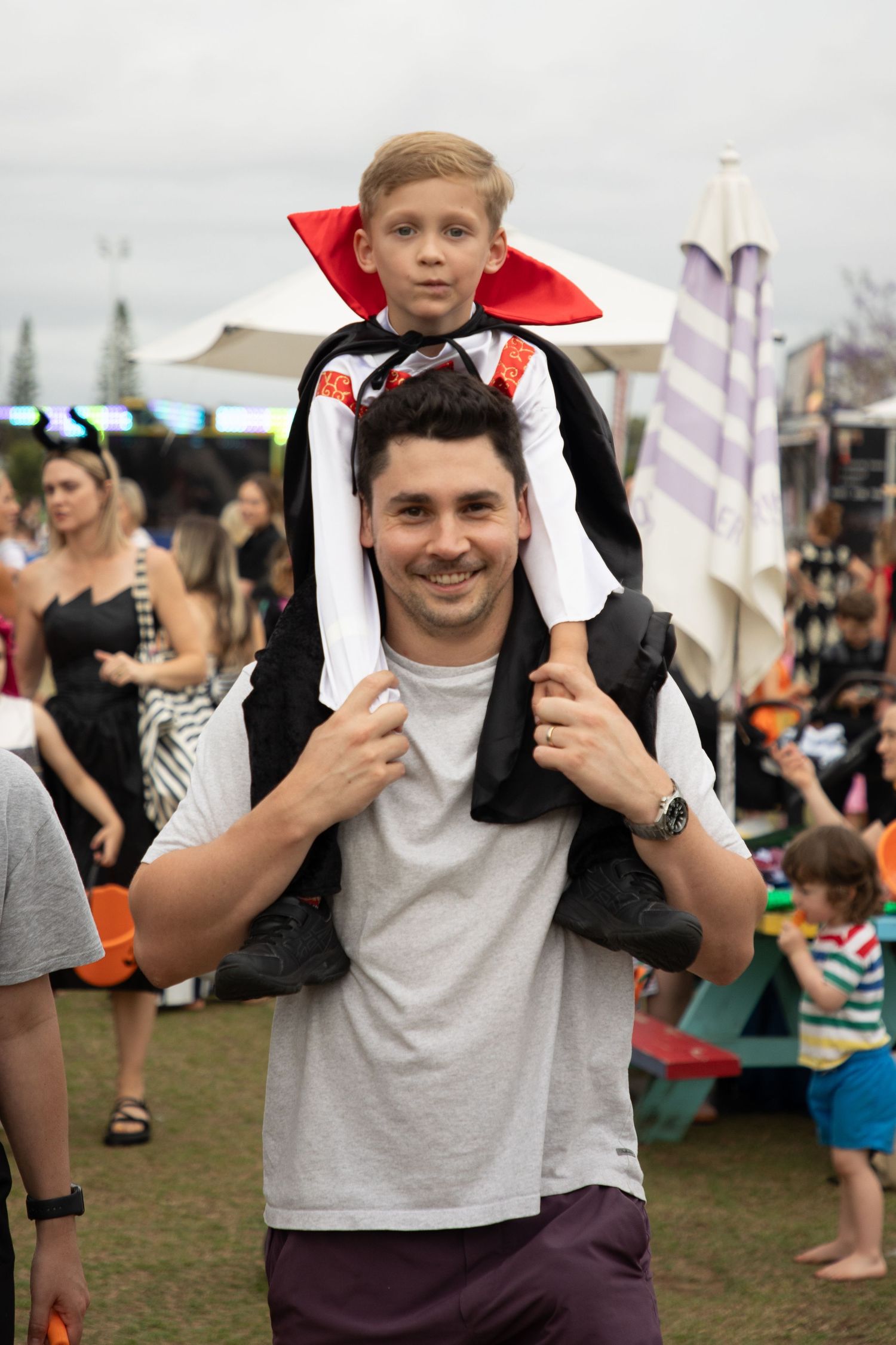 A smiling man carries a young boy dressed in a Dracula Halloween costume on his shoulders outdoors.