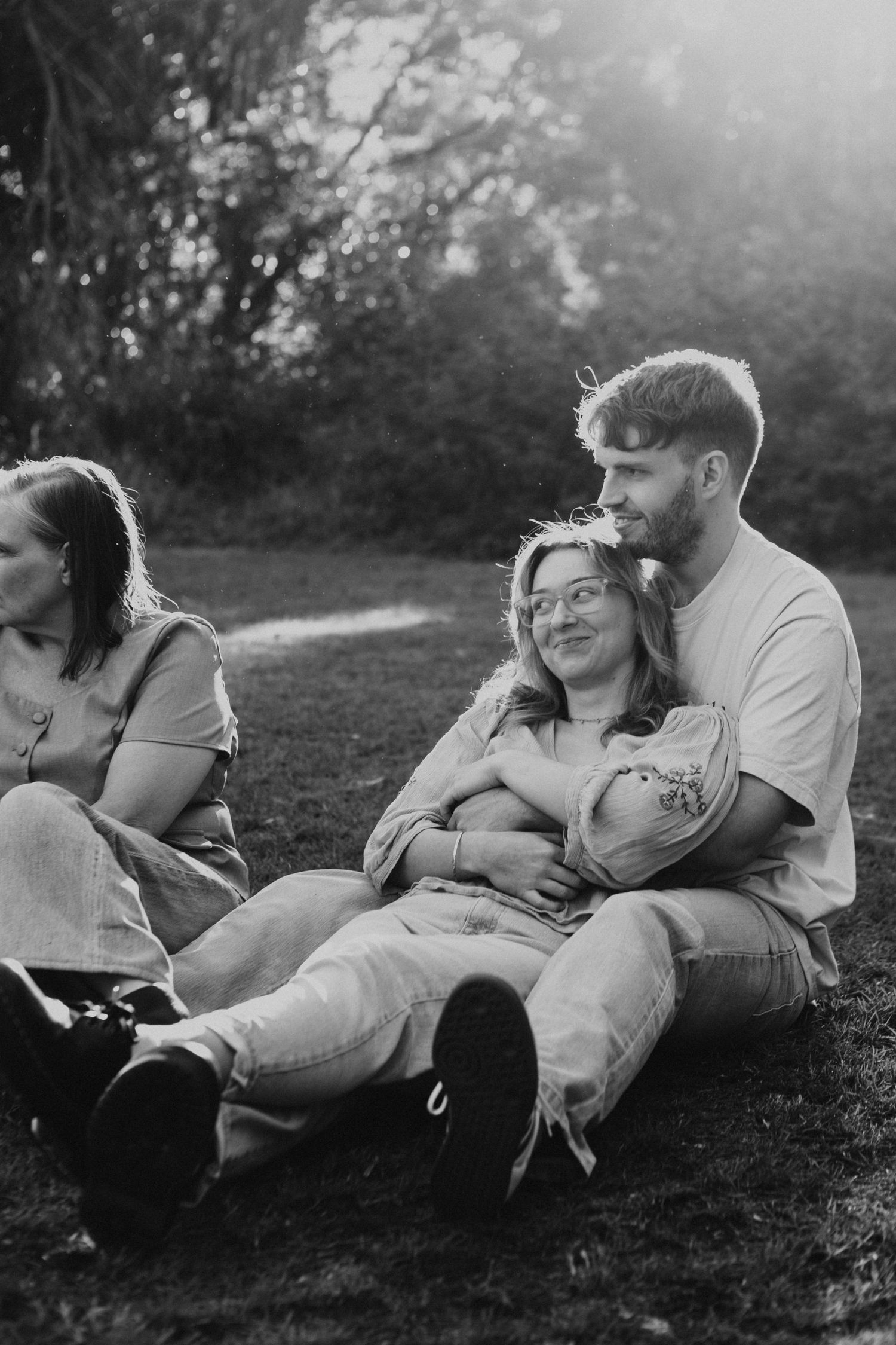 Black and white photo of a couple sitting together on grass in a park, with the man embracing the woman from behind.