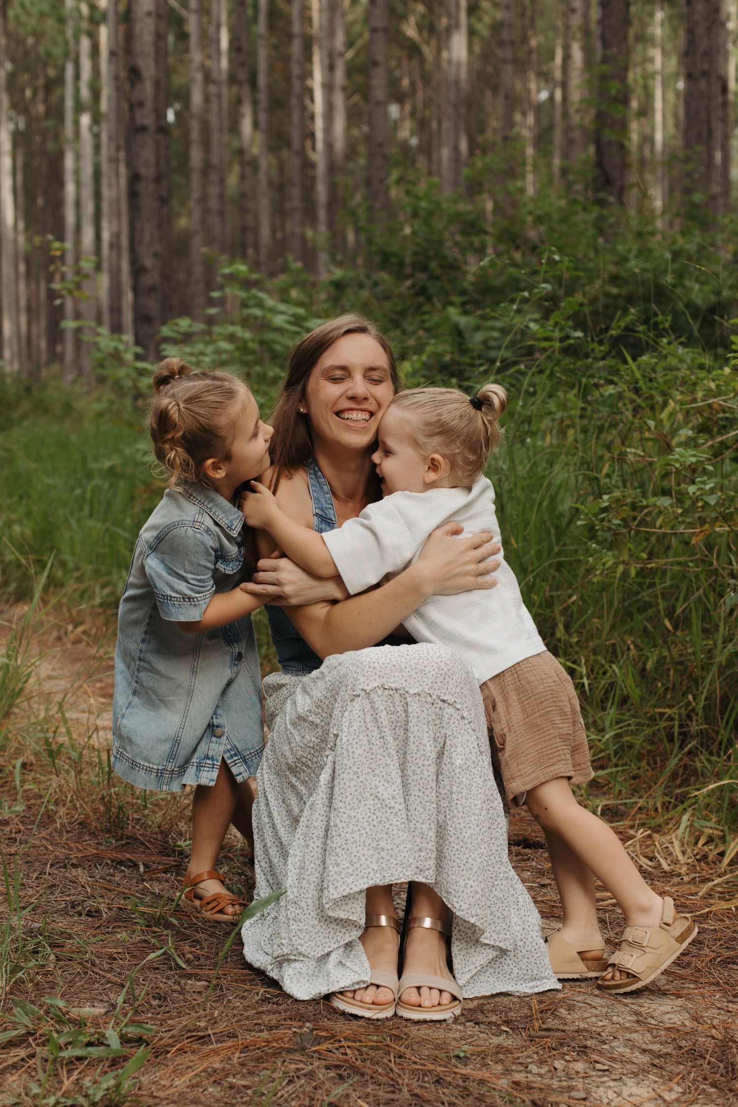 A laughing mother is joyfully embraced by her two young children on a pine forest path.