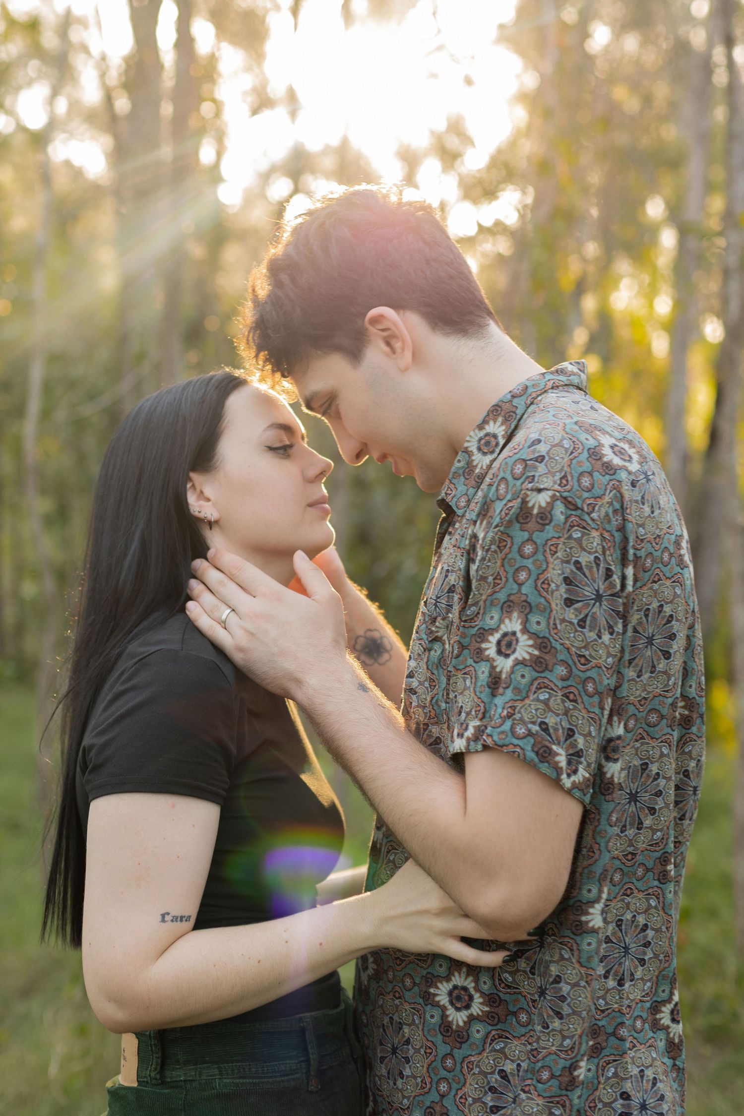 Couple sharing a romantic moment in a sunlit forest, man tenderly holding womans face as they lean in close.