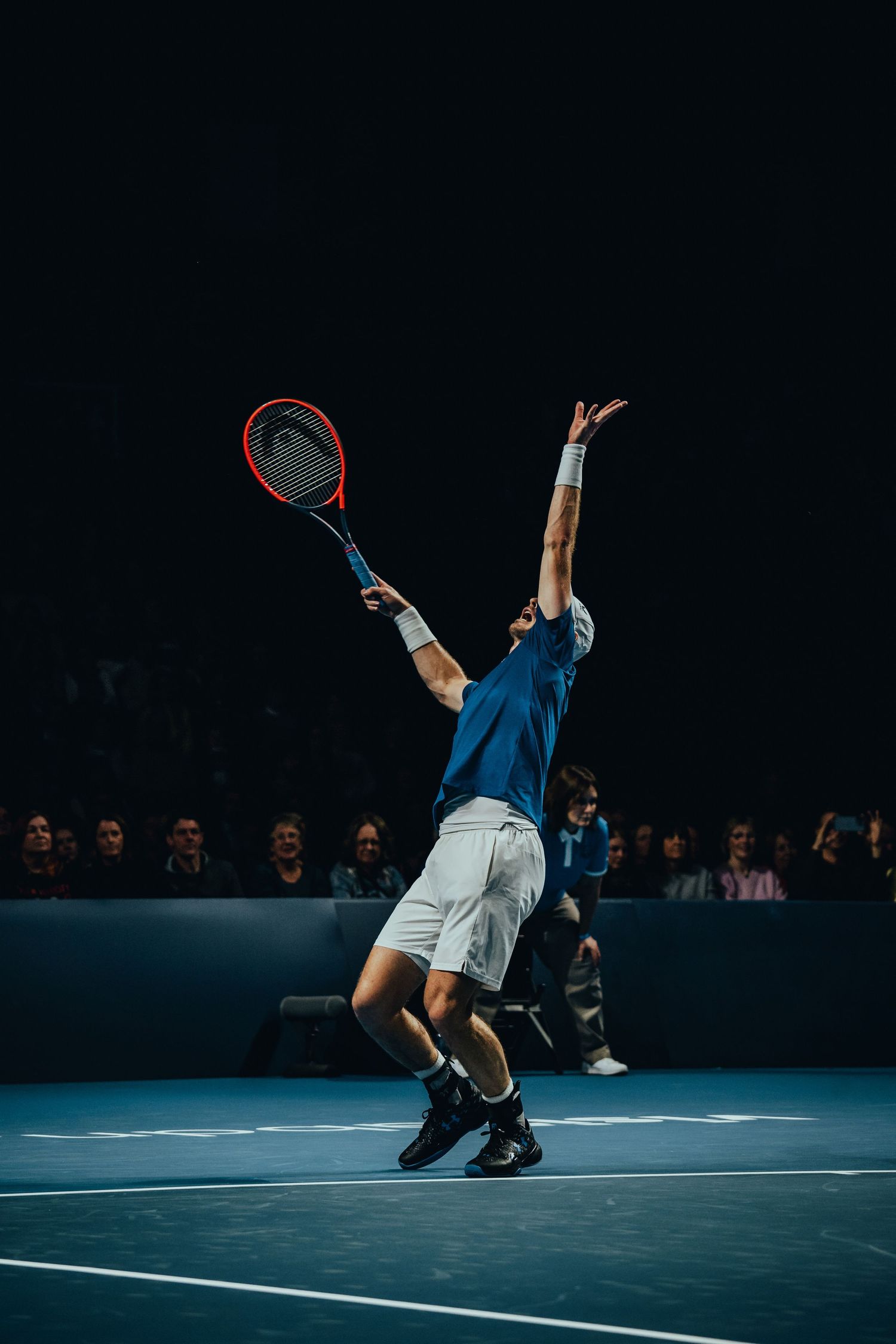 Tennis player in blue shirt serving with a HEAD racket on an indoor court before a crowd.
