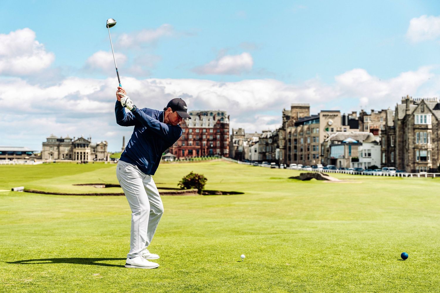 A golfer swings a club at the historic St Andrews golf course on a sunny day with blue skies.