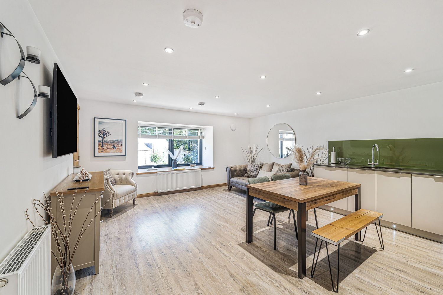 Bright open-plan living and dining space with wooden table, white kitchen units, and large garden-facing window.