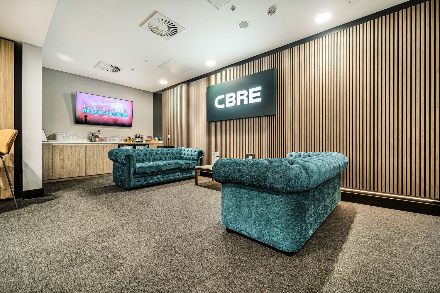 Modern CBRE office lounge with teal velvet Chesterfield sofas, slatted wood feature wall, and wall-mounted TV.