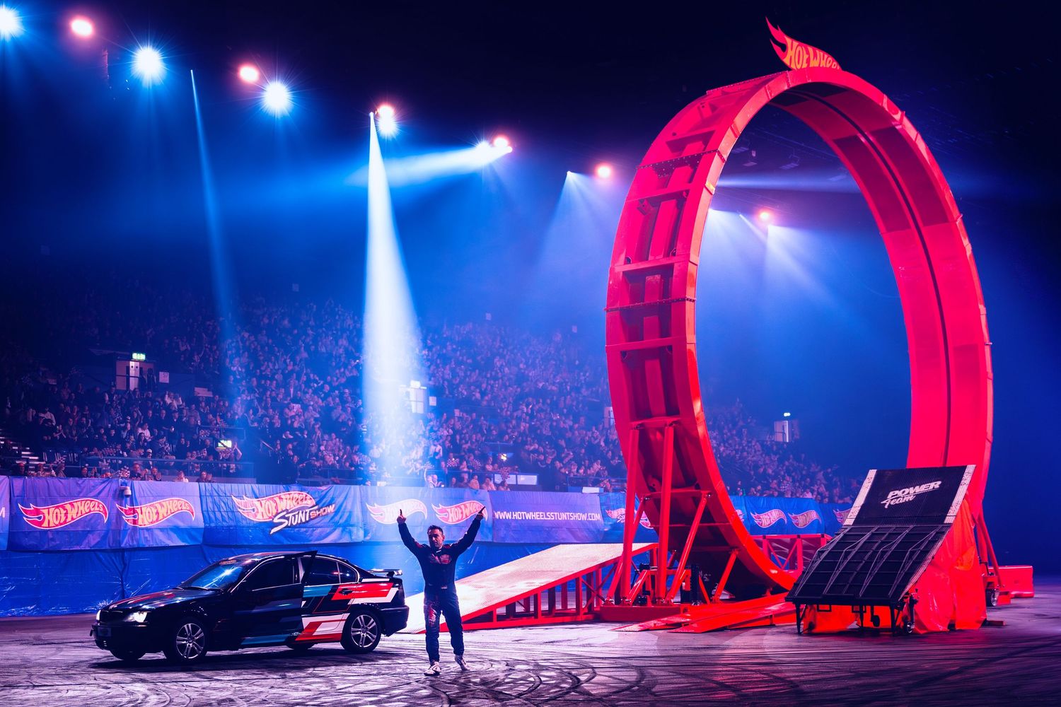 Performer stands triumphantly before a massive red Hot Wheels loop-the-loop in a packed arena.