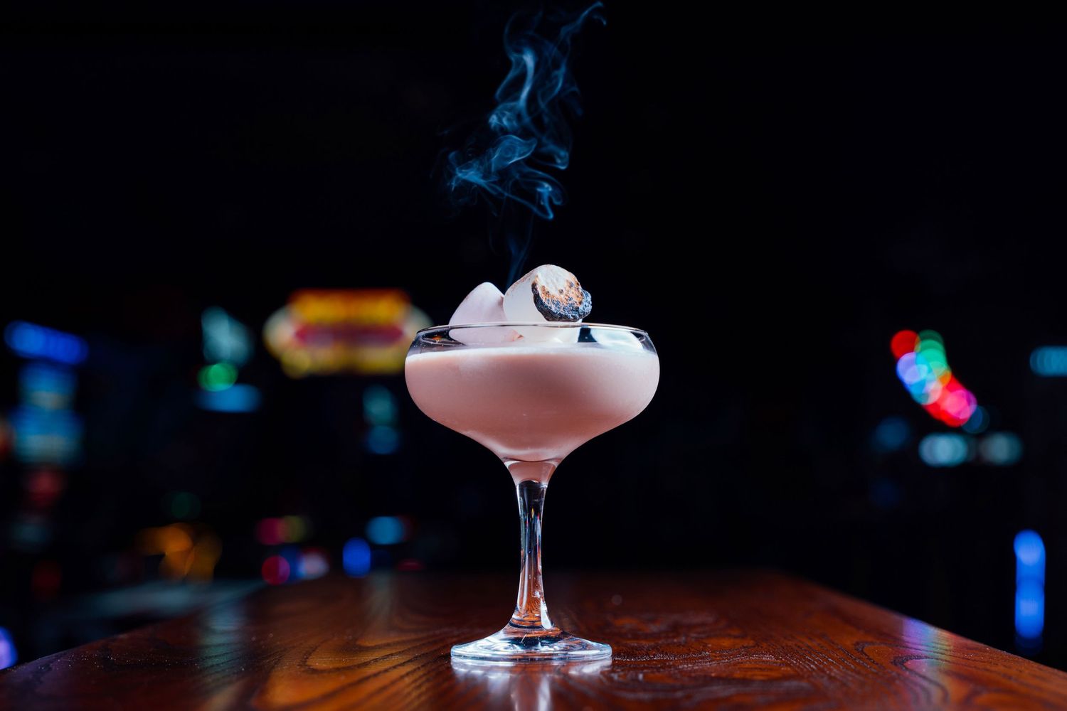 Sophisticated white cocktail with rising smoke in coupe glass against dark bokeh backdrop.