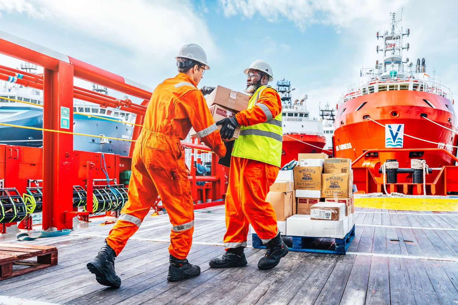 Two crew members in orange coveralls unload cargo boxes on an offshore supply ship deck.