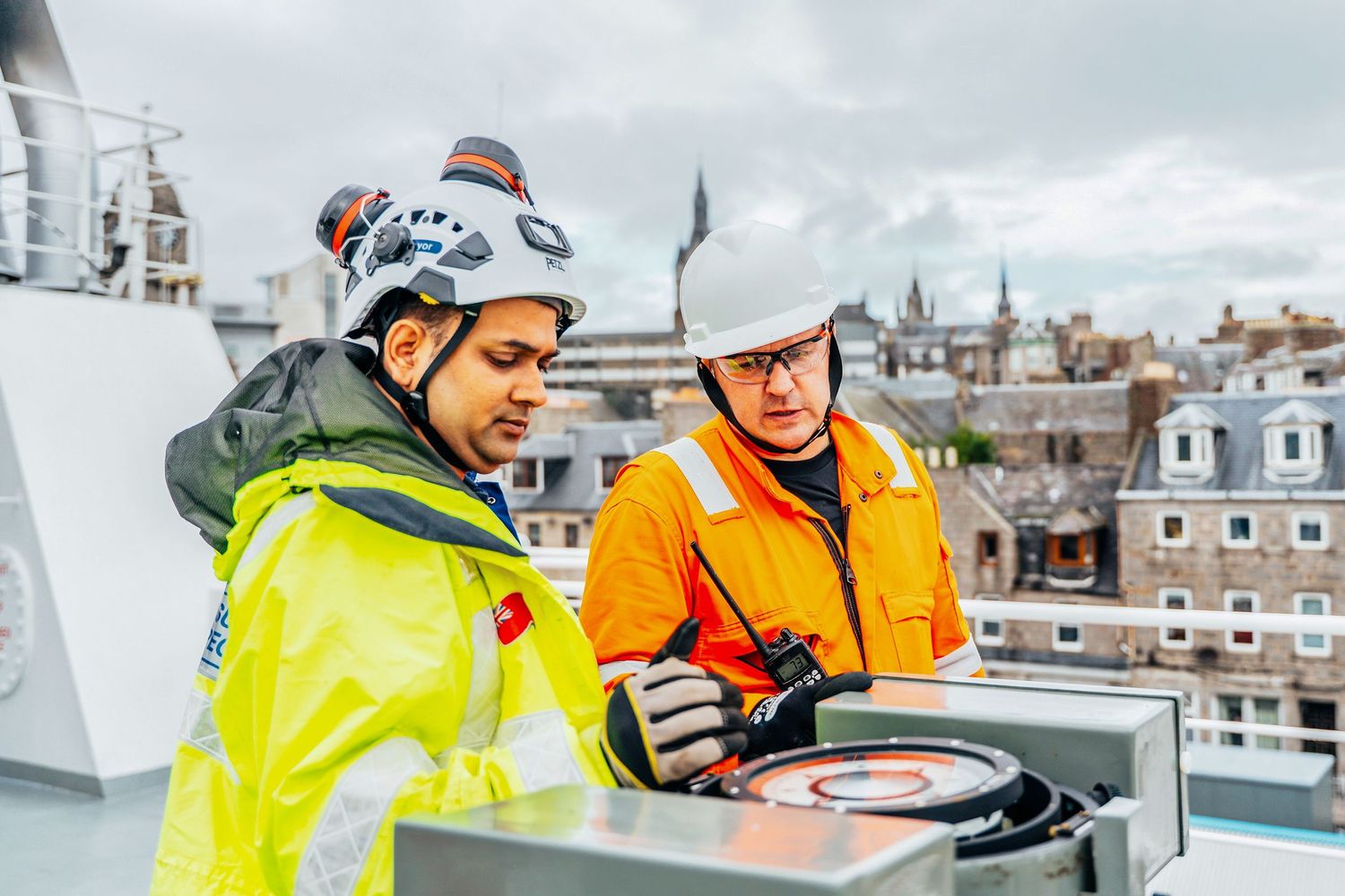 Two engineers in safety gear inspecting rooftop equipment on a building with city views behind them.