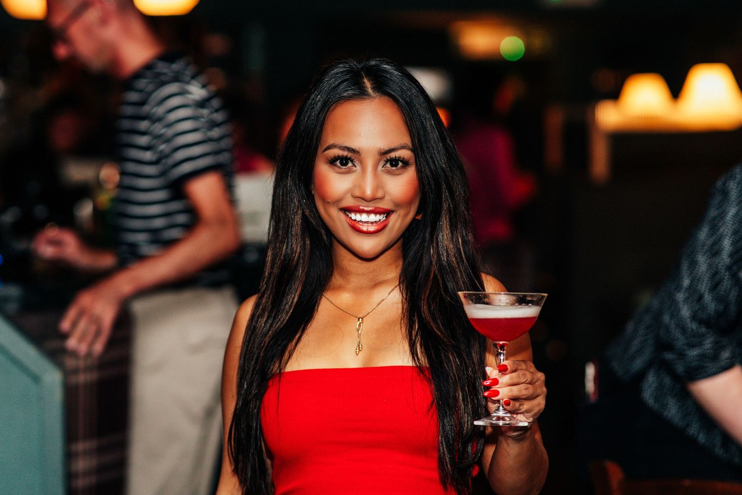 Smiling woman in red dress holding a martini cocktail at a lively bar or nightclub event.