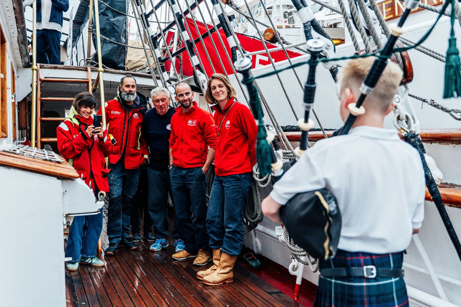 Group of people in red jackets posing on a tall ship deck with a bagpiper.