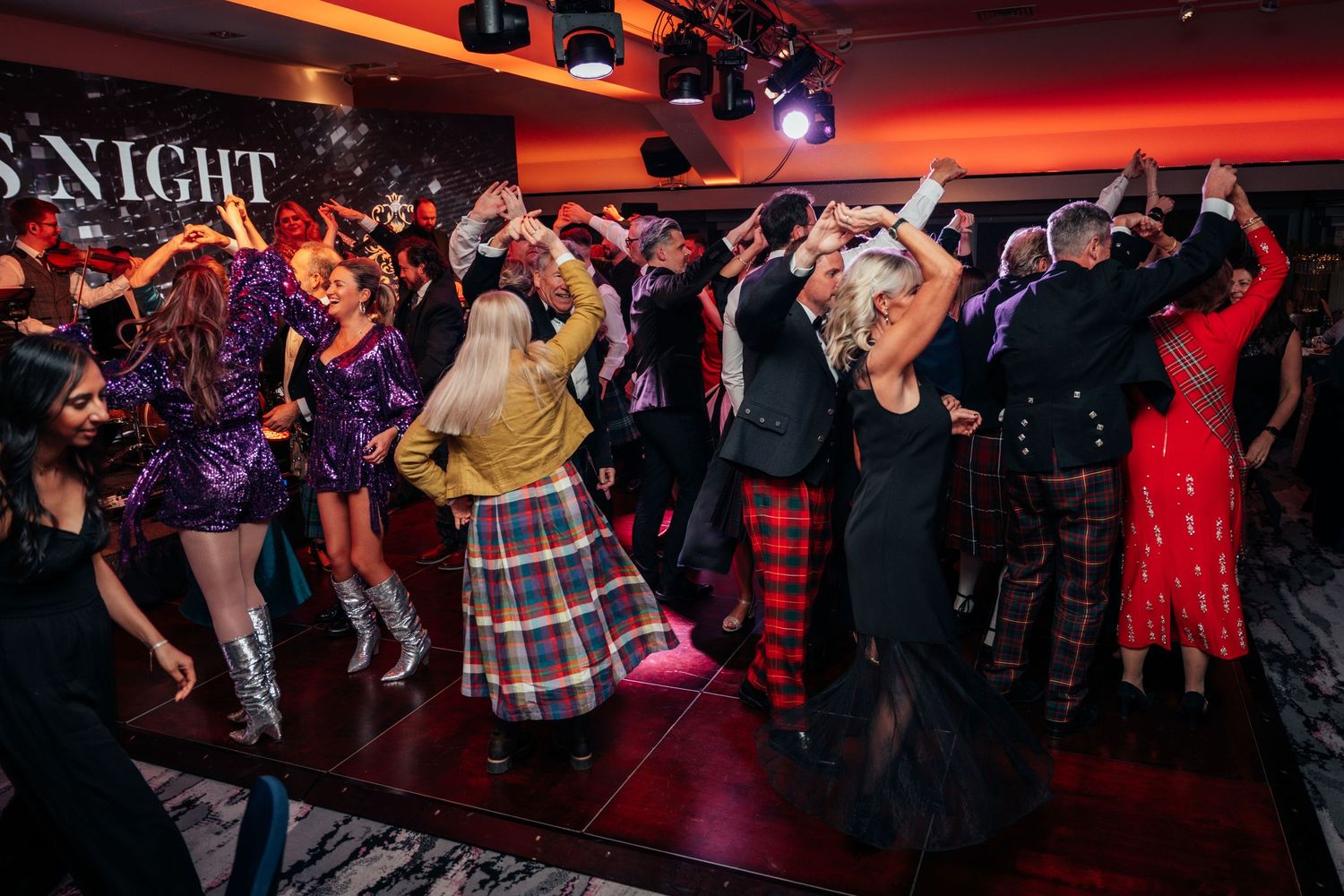 Guests in formal and Scottish tartan attire dance energetically at a lively Burns Night celebration event.