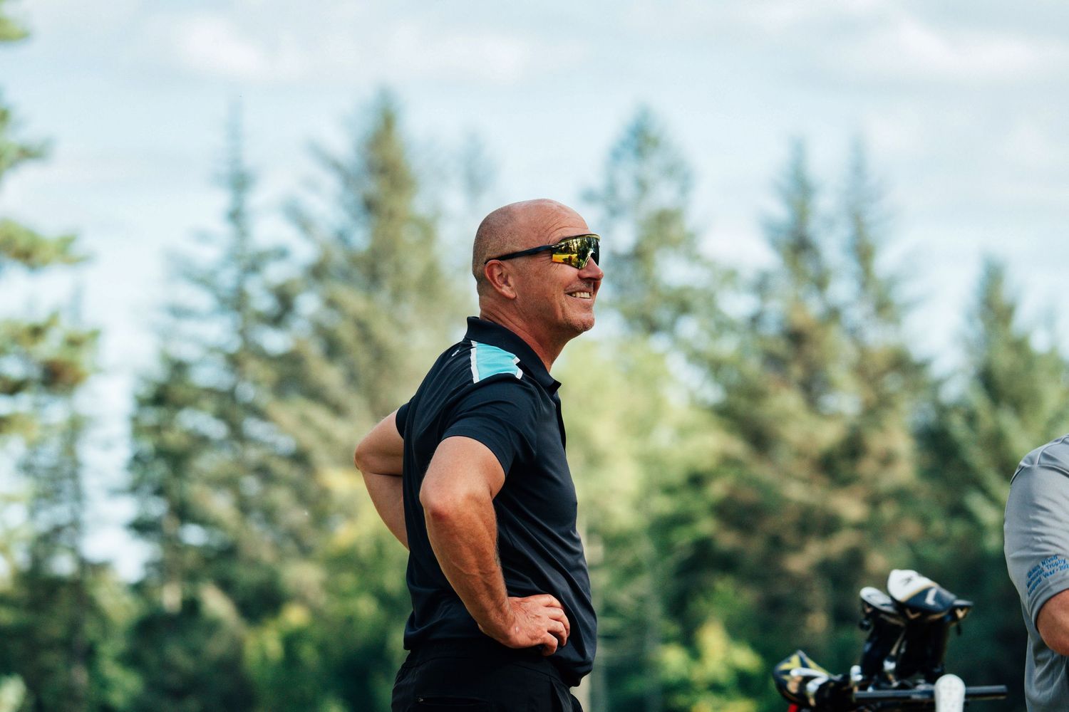 A bald man in sunglasses smiles outdoors with tall pine trees in the background.