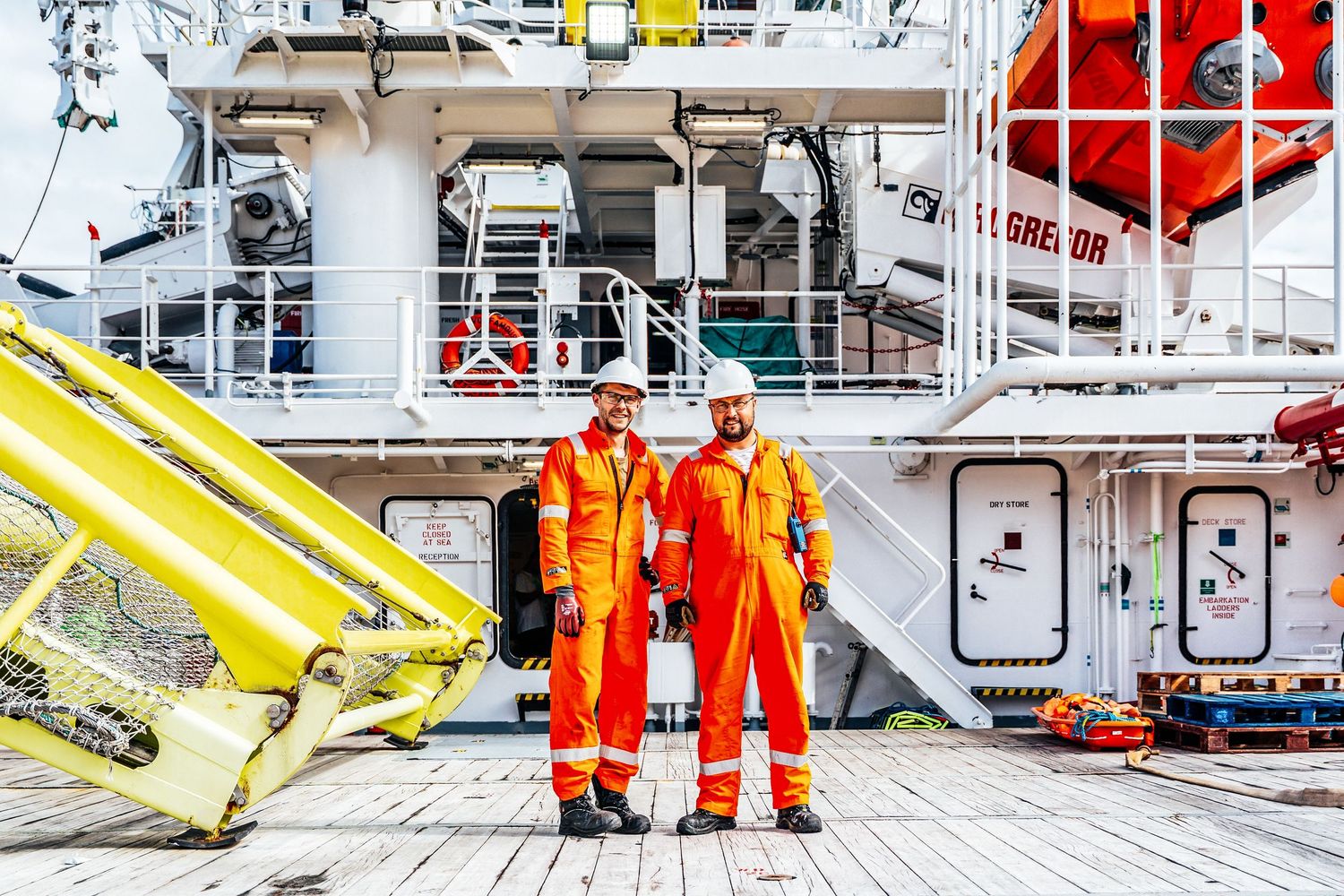 Two workers in orange coveralls and hard hats pose on an offshore vessel deck with yellow equipment.