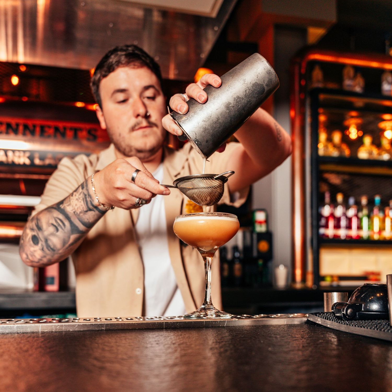 Tattooed bartender straining a cocktail into an elegant coupe glass at a well-stocked bar.