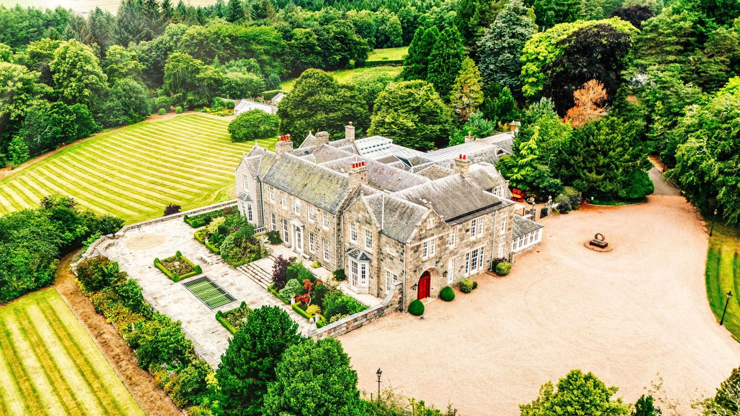 Aerial view of a stone manor house with formal gardens, gravel driveway, and surrounding woodland landscape.