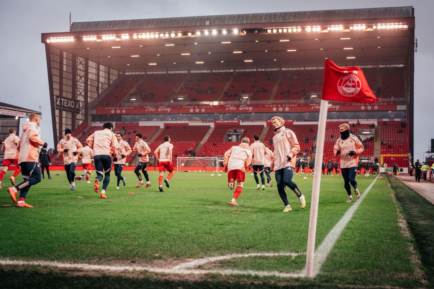 Football players warming up on a floodlit red stadium pitch near a corner flag before a match.