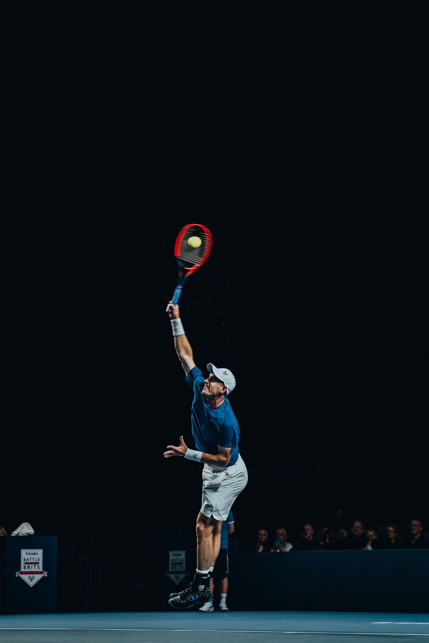 Tennis player leaping to serve with a HEAD racket under dramatic lighting on an indoor court.