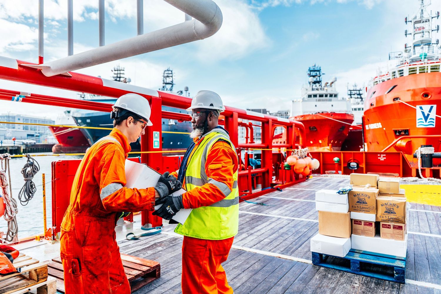 Two offshore workers in safety gear review documents on a supply vessel deck in port.