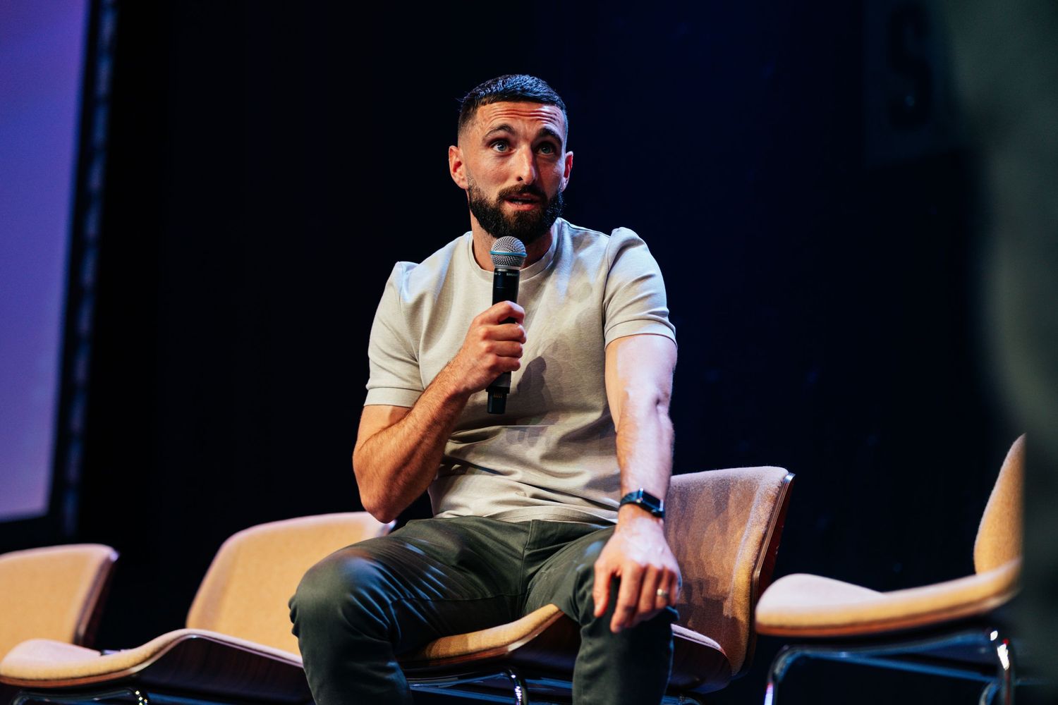 Bearded man in grey t-shirt speaking into a microphone while seated on stage at an event.