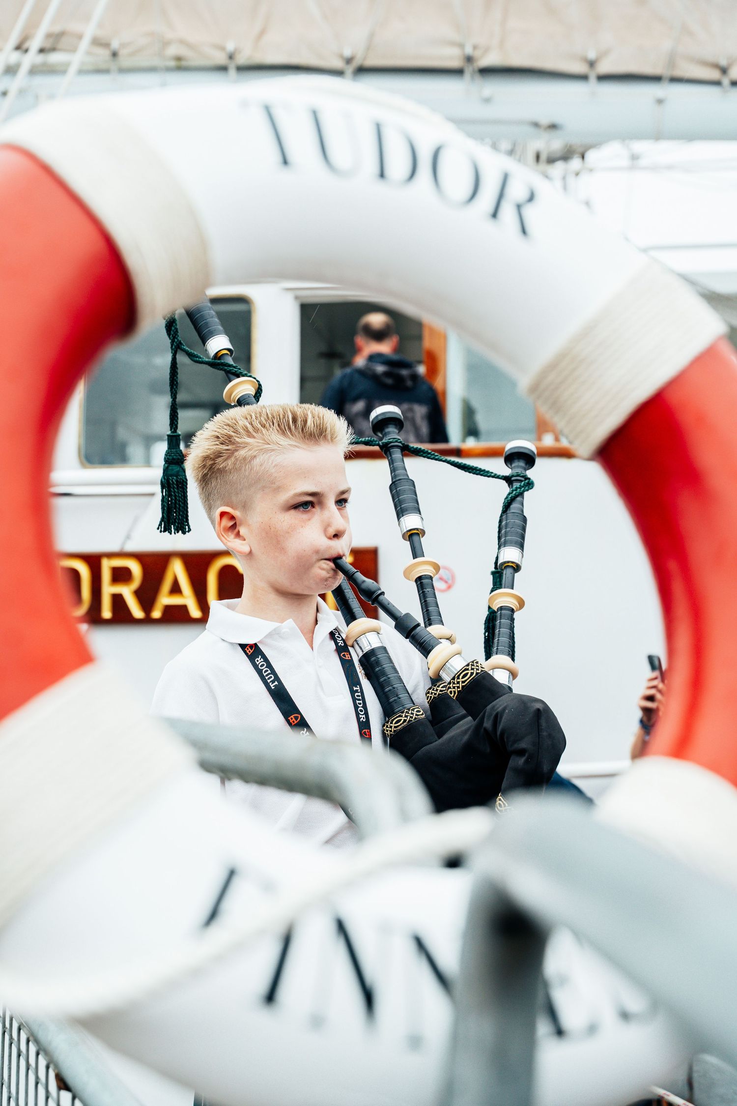 Young boy playing bagpipes framed by a Tudor life preserver ring on a ship deck.