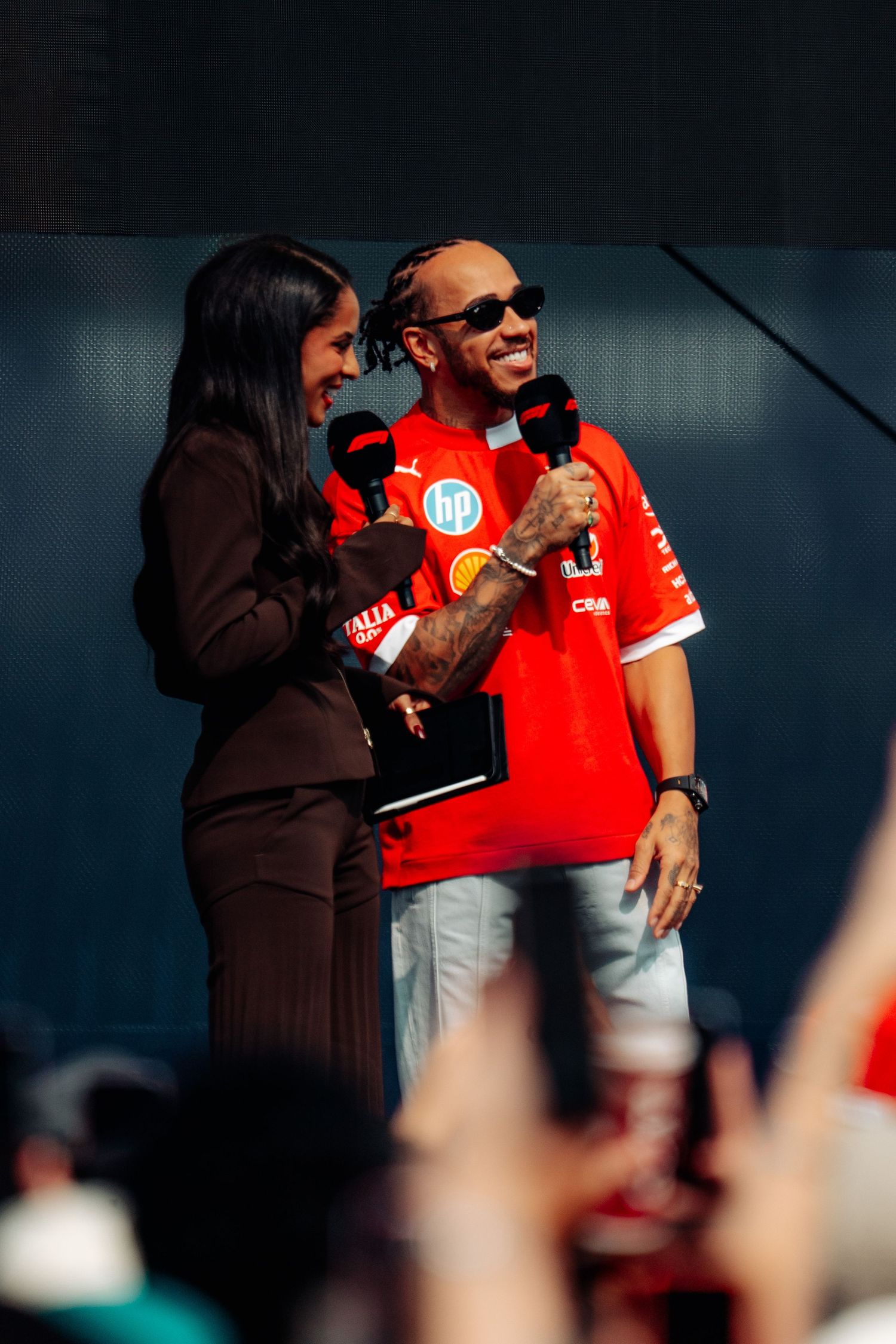 Lewis Hamilton in Ferrari kit laughs with female interviewer on stage at a Formula 1 fan event.