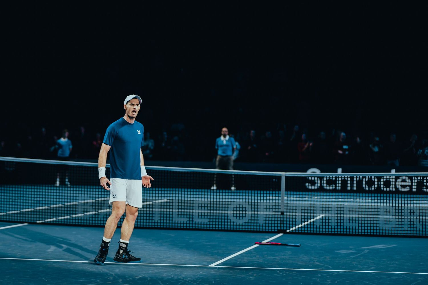 Tennis player standing near the net on an indoor court at a Schroders sponsored match.