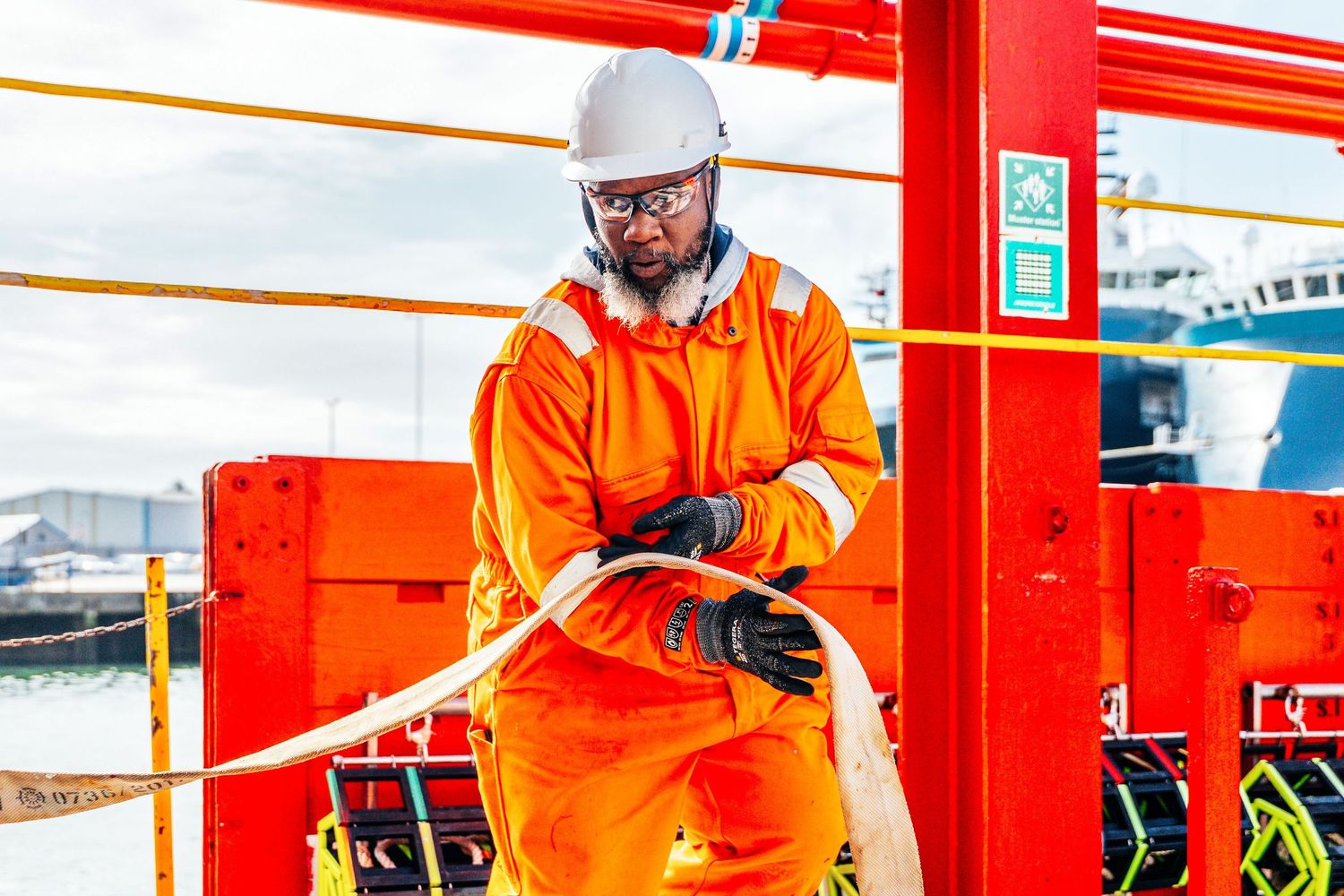 Offshore worker in orange coveralls handles mooring rope on a supply vessel at port.
