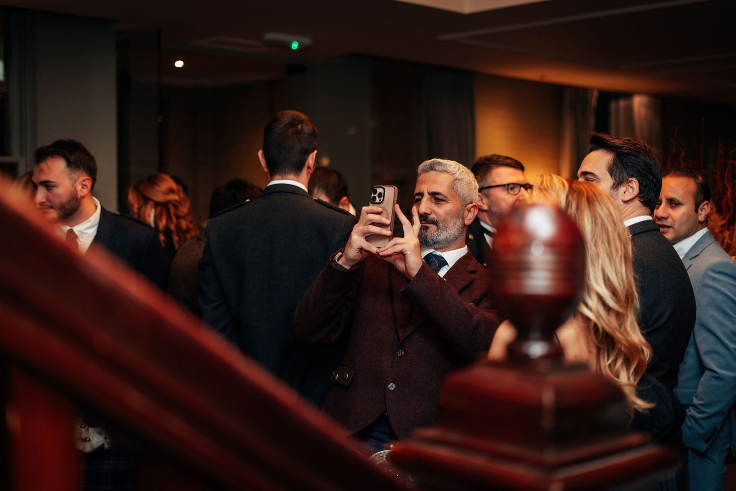 Man in suit taking selfie at crowded upscale indoor social gathering event.